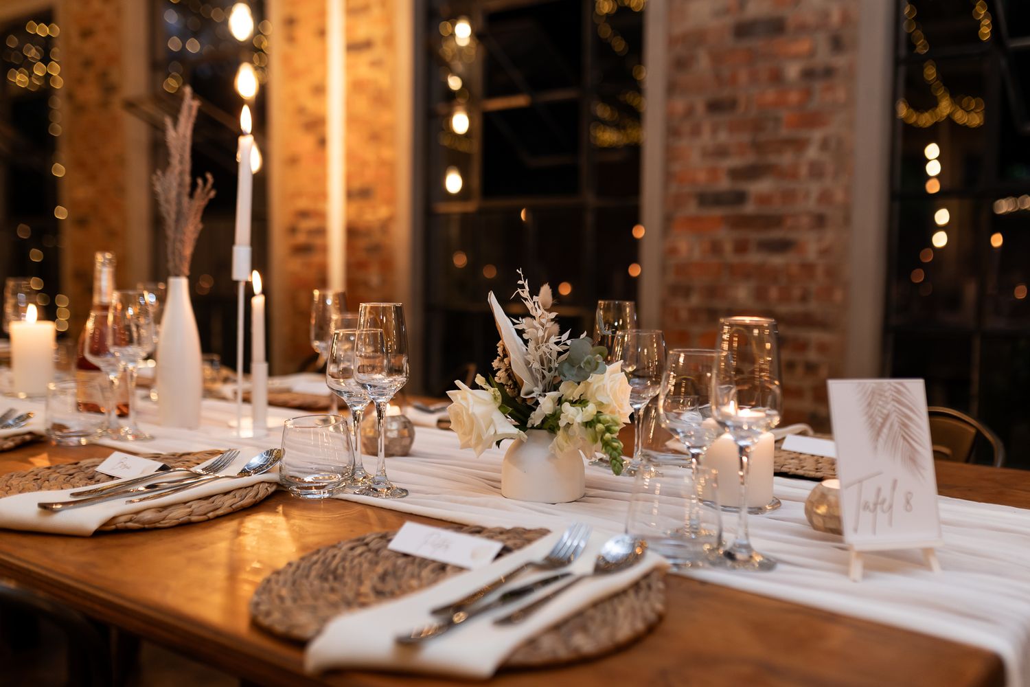 Elegant wedding reception table setting with white linens and floral centerpieces in rustic brick venue.