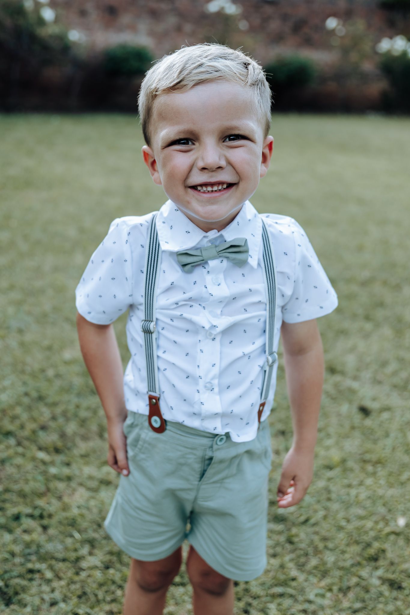 Young ring bearer in mint green shorts and suspenders smiles at camera.