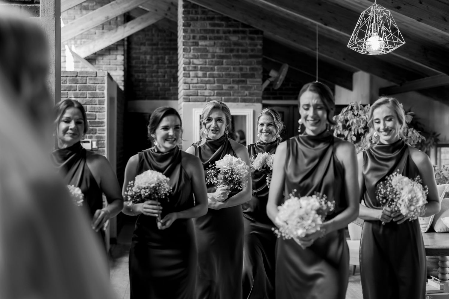 Black and white photo of bridesmaids in dark dresses holding bouquets during wedding ceremony.