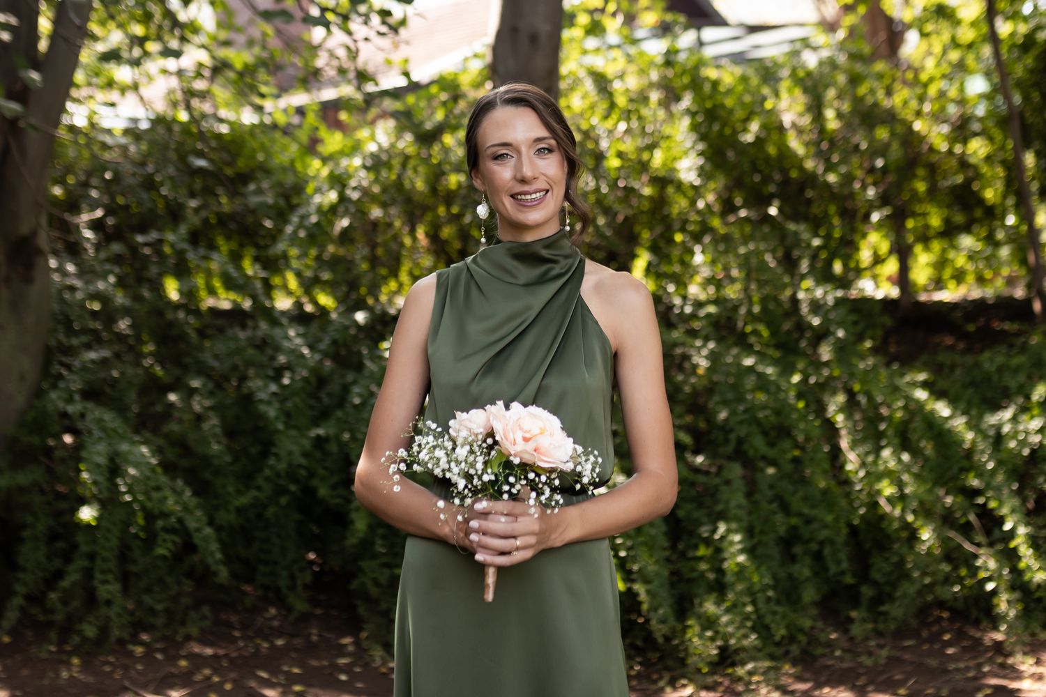 A person in a sage green halter dress holds a pink bouquet standing outdoors in a garden setting.