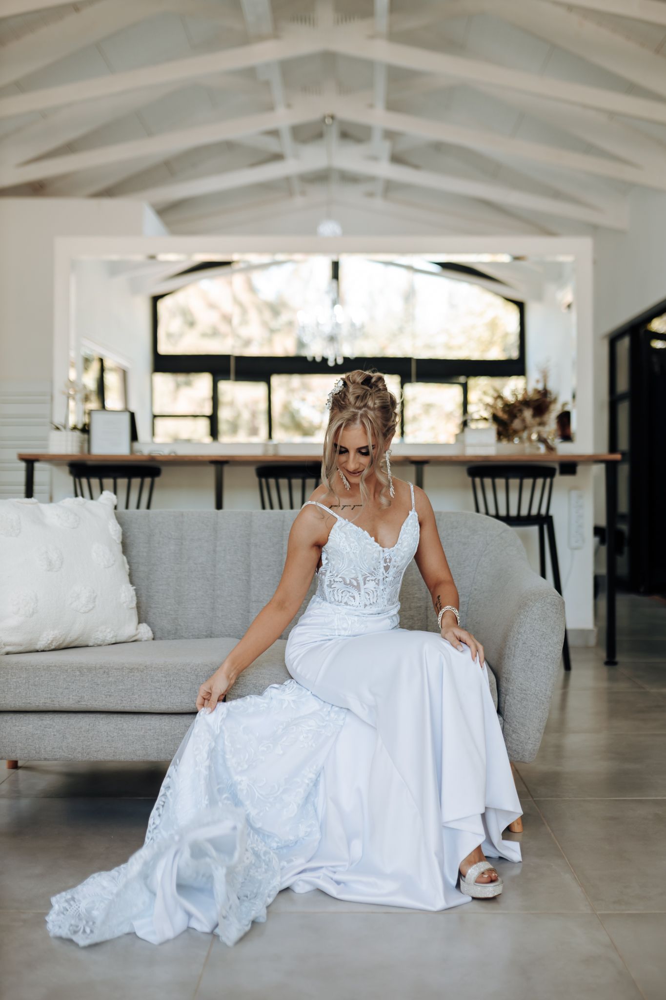Person in elegant white wedding dress sits gracefully on gray sofa in modern room with vaulted ceiling.