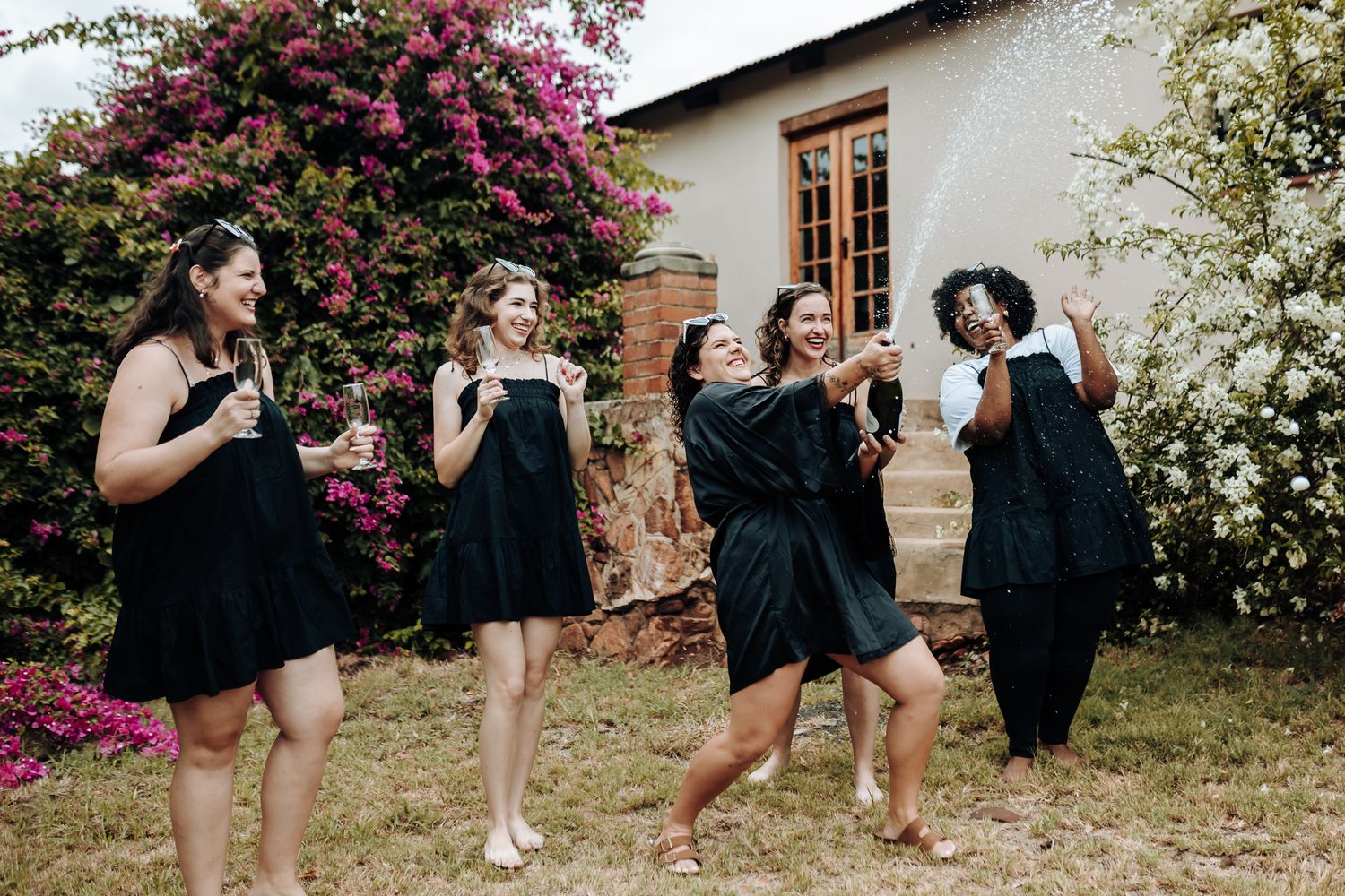 Group in matching black dresses celebrating in garden with blooming bougainvillea and white house backdrop.