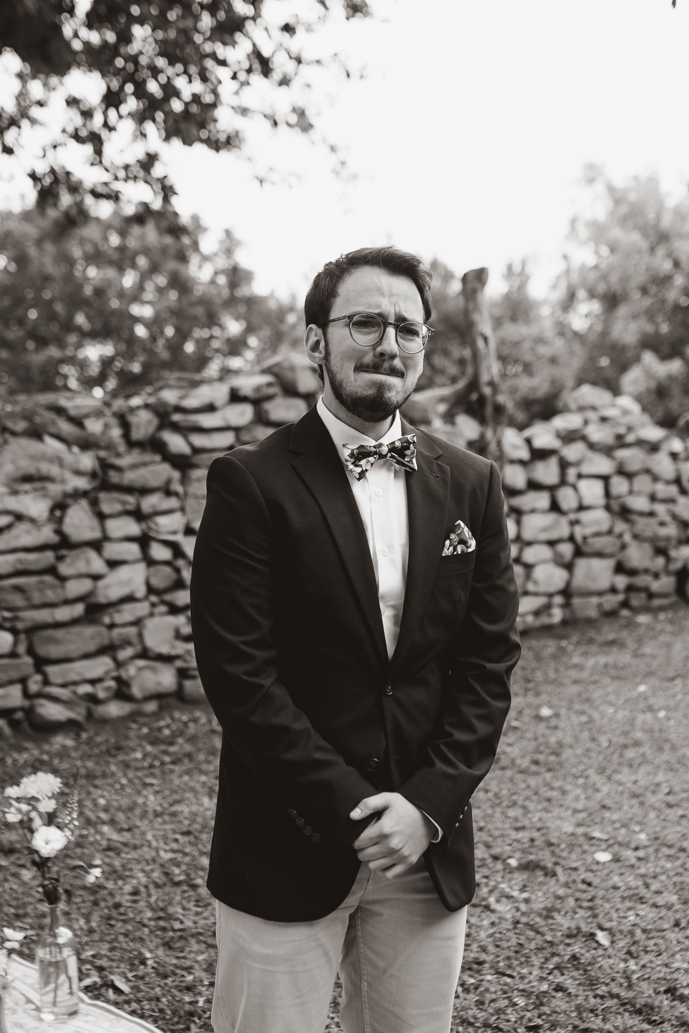 Black and white portrait of person in formal attire with bow tie against stone wall backdrop.