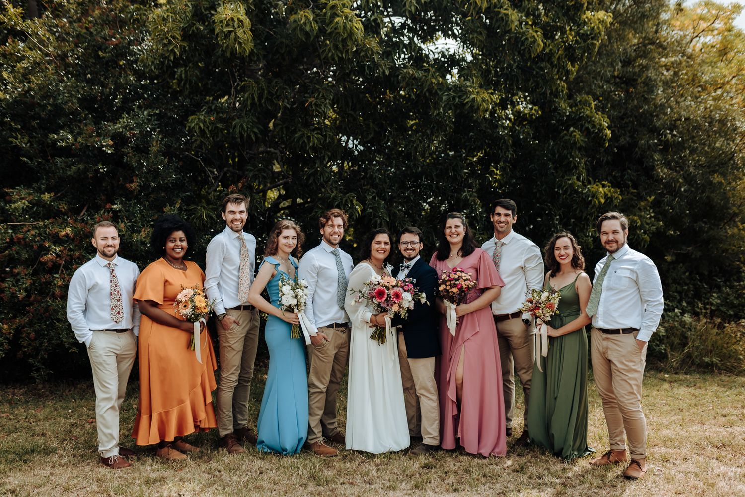 Group wedding photo with bridal party wearing vibrant orange, blue, pink and green dresses outdoors.