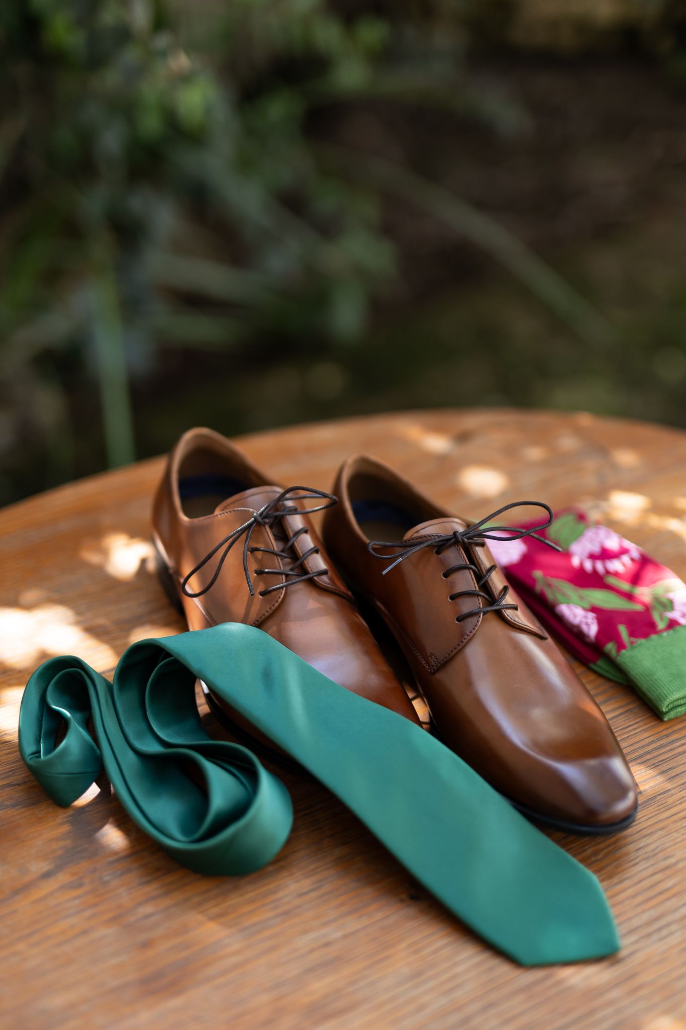 Brown leather dress shoes with emerald green tie and boutonniere on wooden surface.