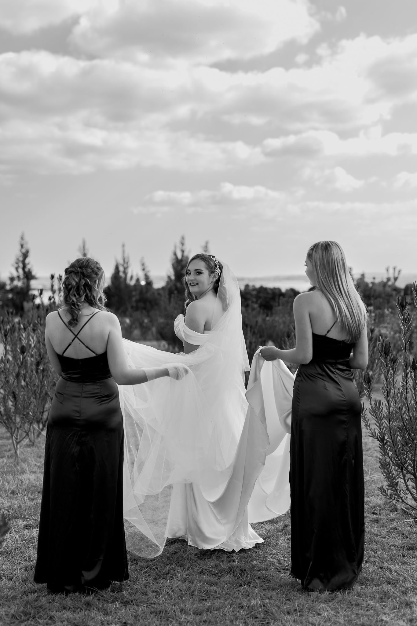 Black and white photo of bridesmaids helping adjust a wedding dress train in an outdoor field setting.