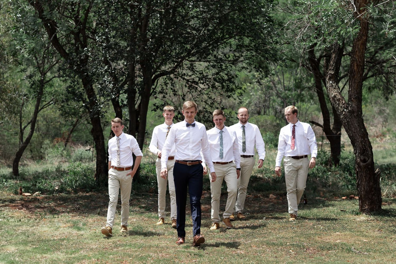 Groomsmen in coordinated white shirts and khakis walk together through wooded outdoor setting.