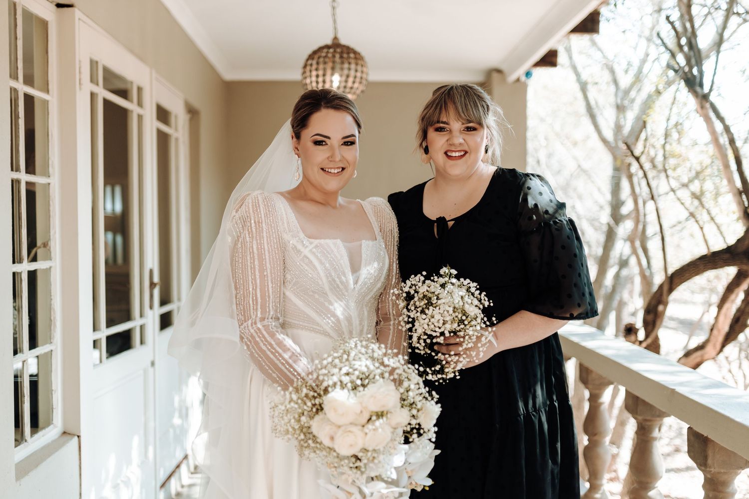 Bride and bridesmaid smile while holding white floral bouquets on covered porch.