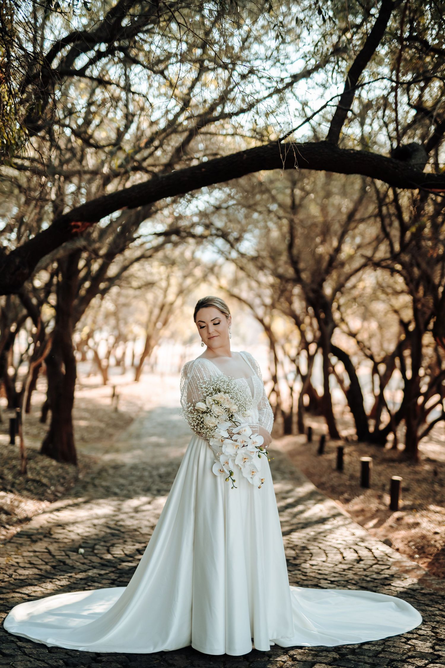 Elegant bride in flowing white dress holds bouquet while standing on stone path under trees.