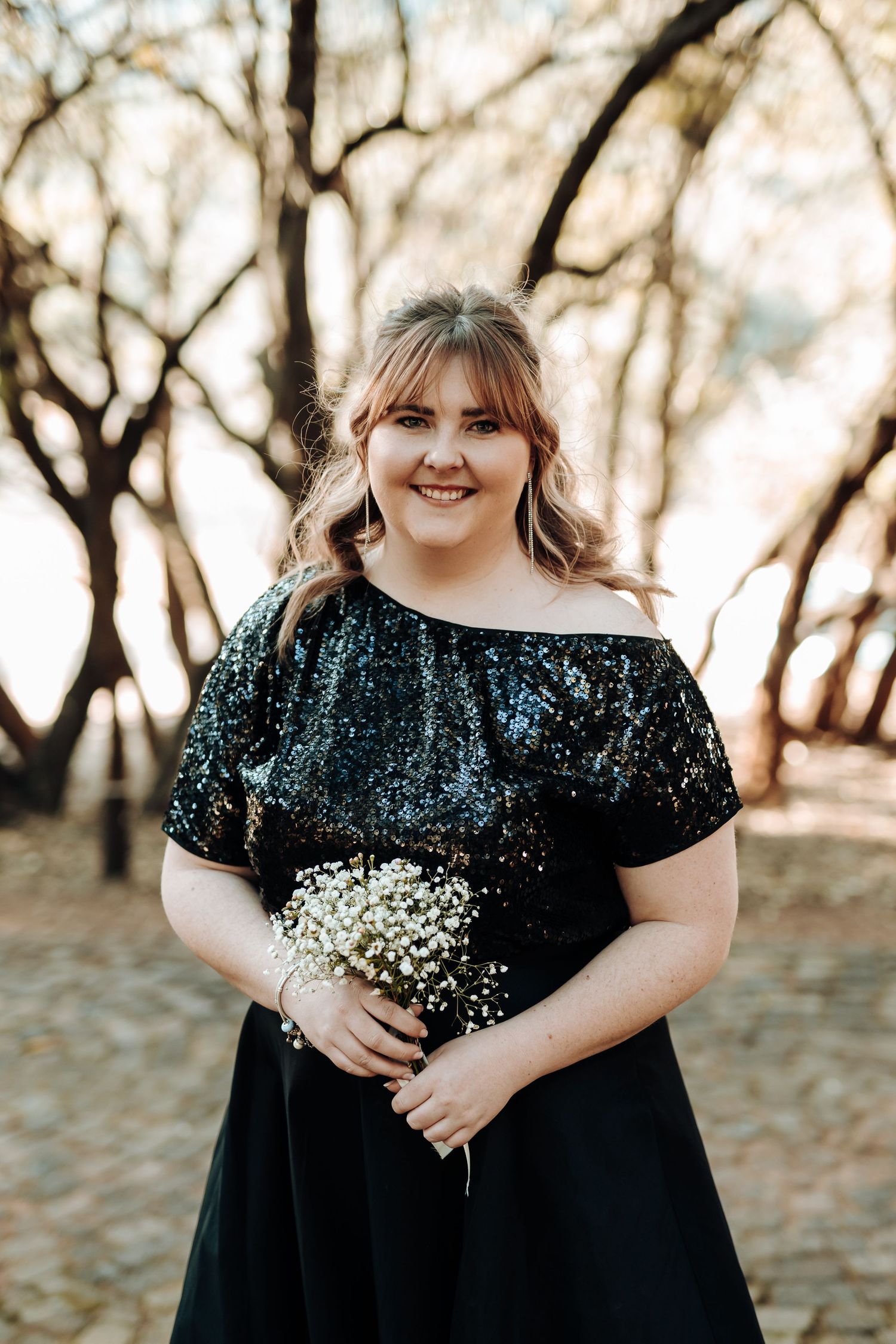 Bridesmaid in off-shoulder sequin black dress holds baby's breath bouquet in autumn setting.