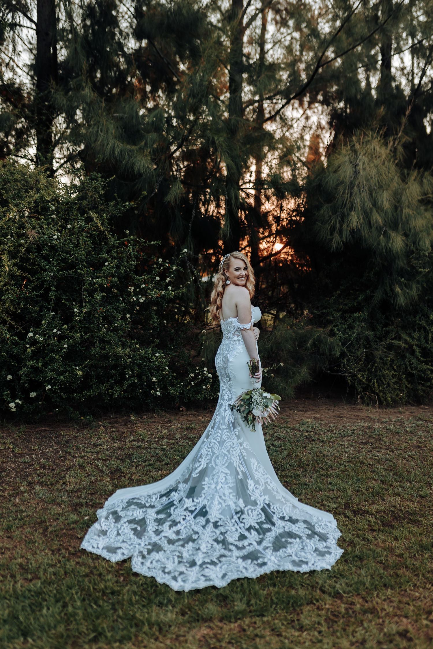 Wedding dress with intricate lace train photographed against evergreen forest at golden hour.