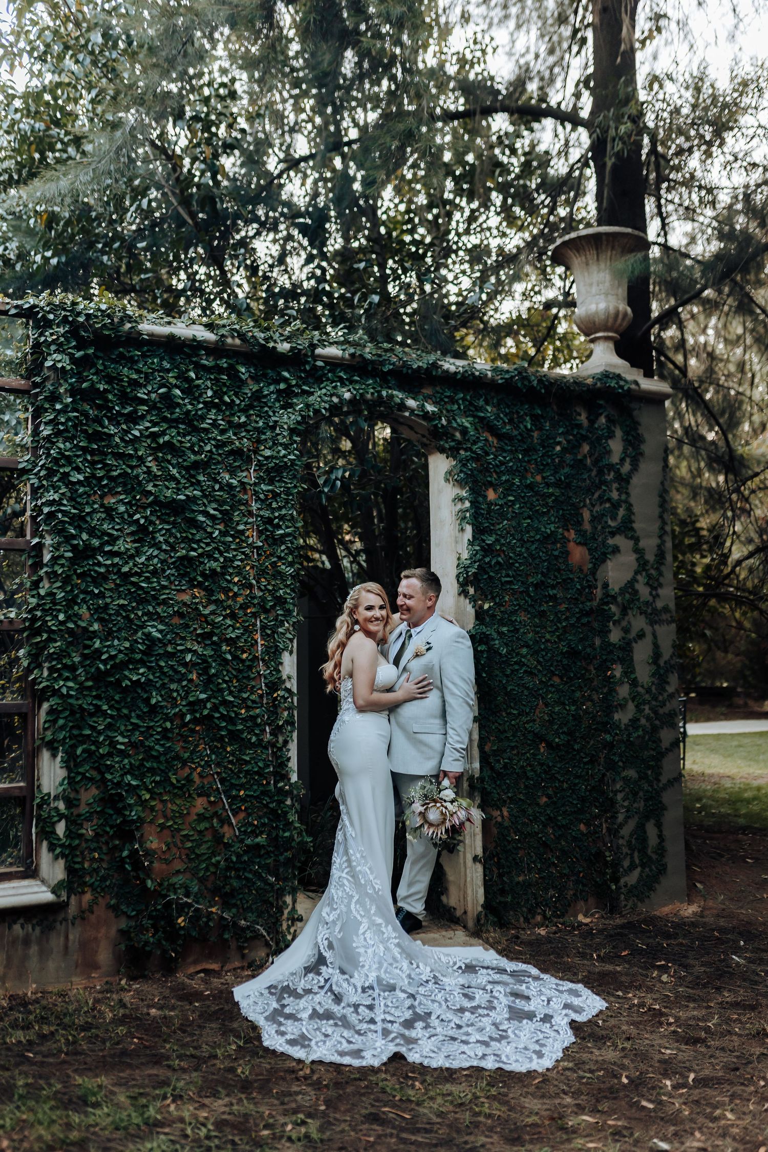 Wedding couple poses against ivy-covered wall with dramatic lace train in garden setting.