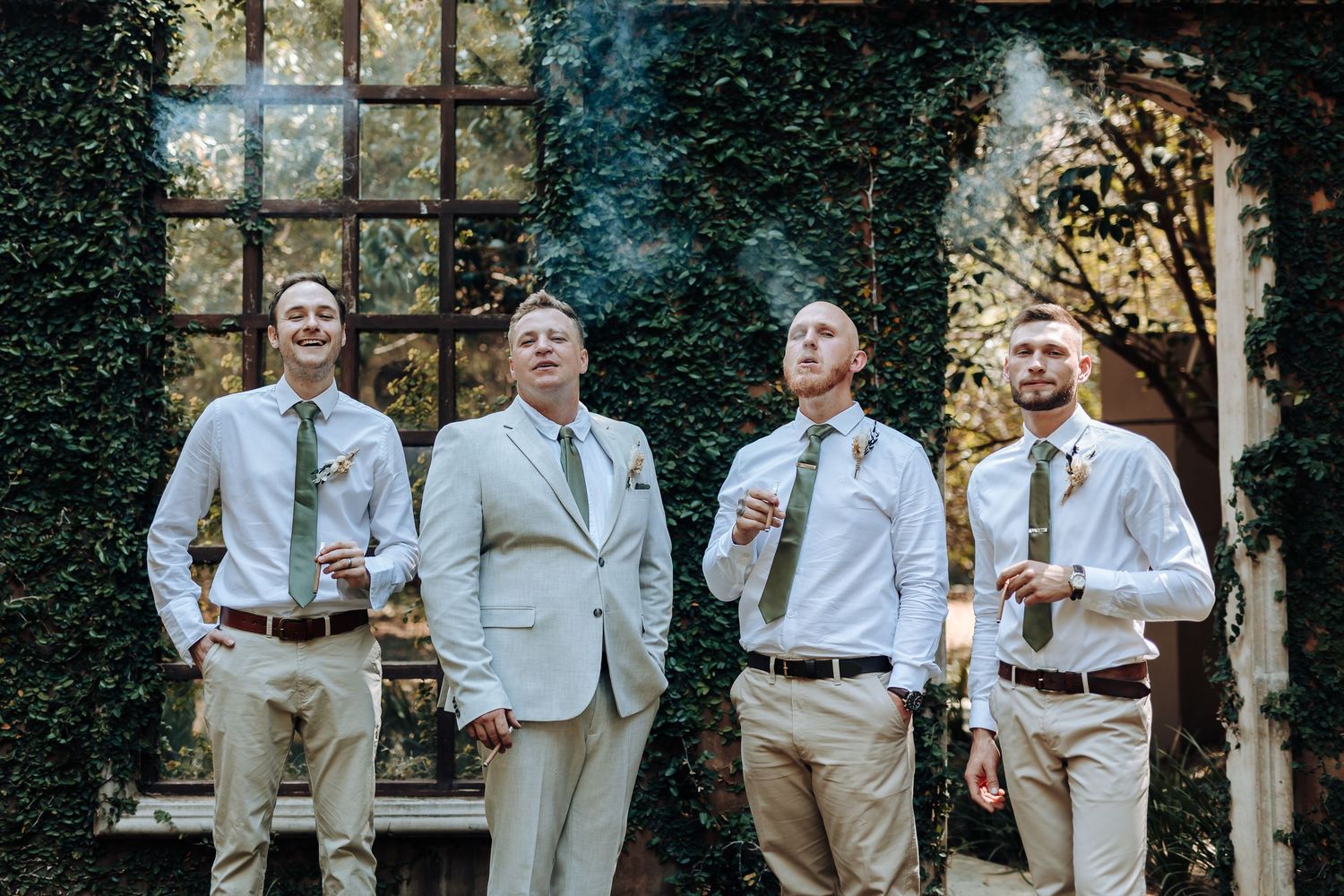 Wedding party shares laughs while posing in light colored attire against stone wall.