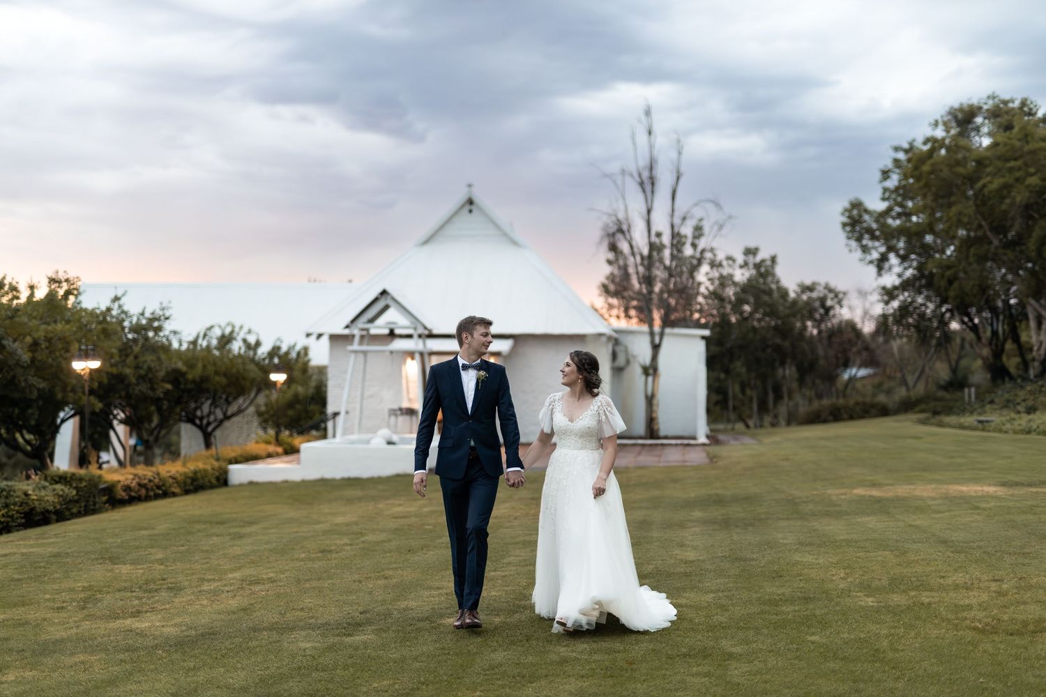 Couple walking hand in hand on green lawn with white chapel in background during sunset.
