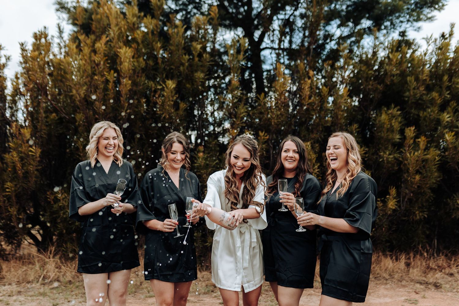 Wedding party popping champagne outdoors in matching black robes.