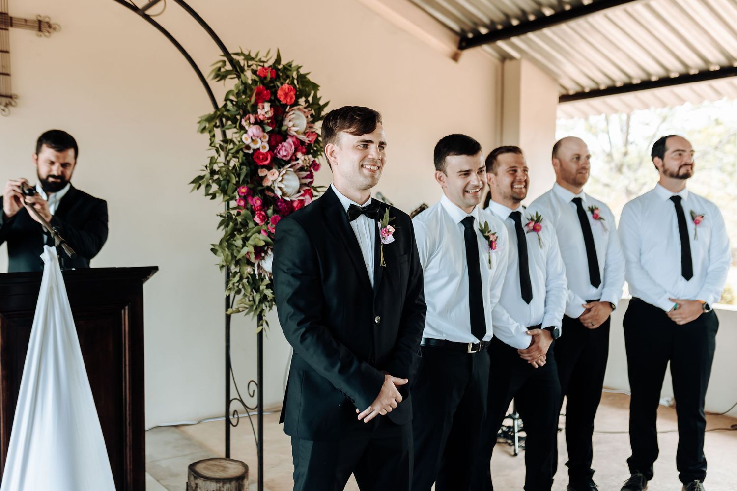 Wedding party stands under floral arch during outdoor ceremony with pink and red flower arrangements.