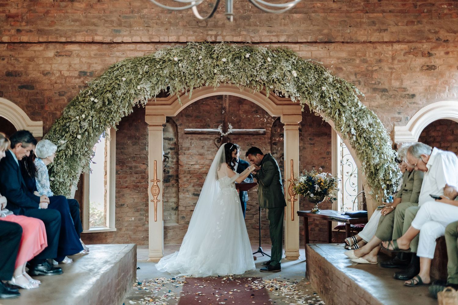 Wedding ceremony under a floral arch in a rustic brick venue with guests seated on both sides.