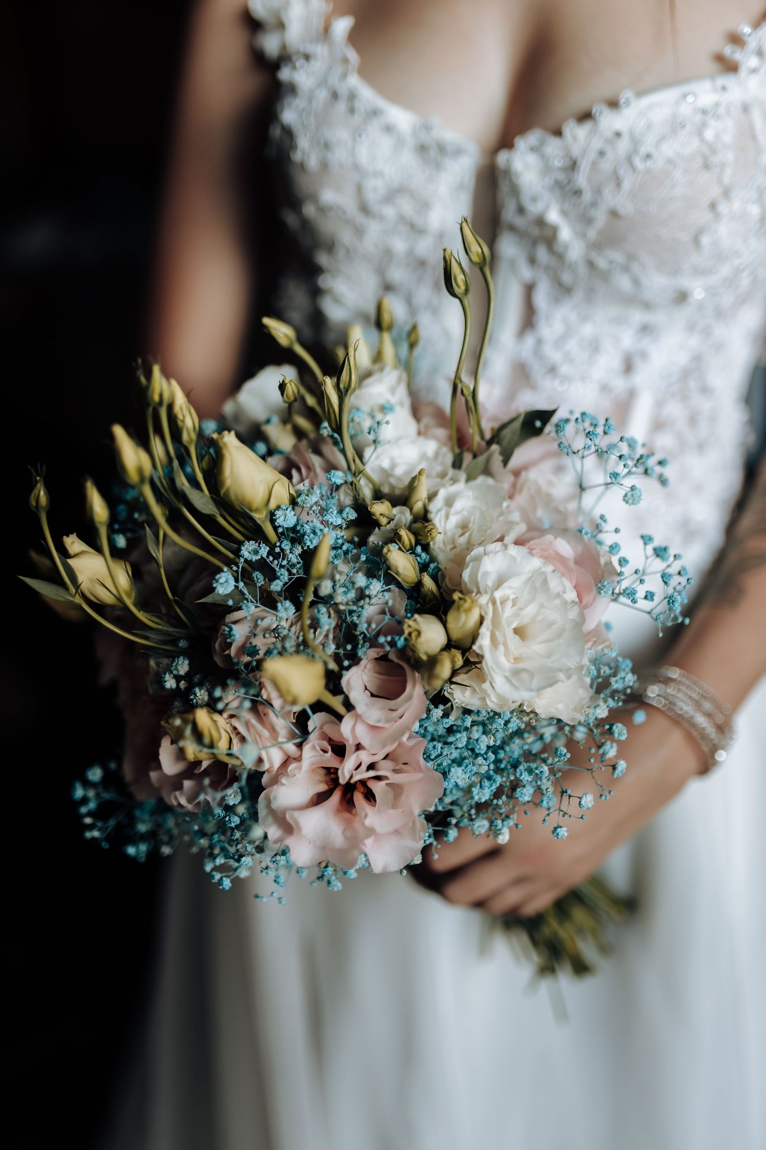 A delicate wedding bouquet featuring white peonies, blue baby's breath, and pink roses against a white lace dress.