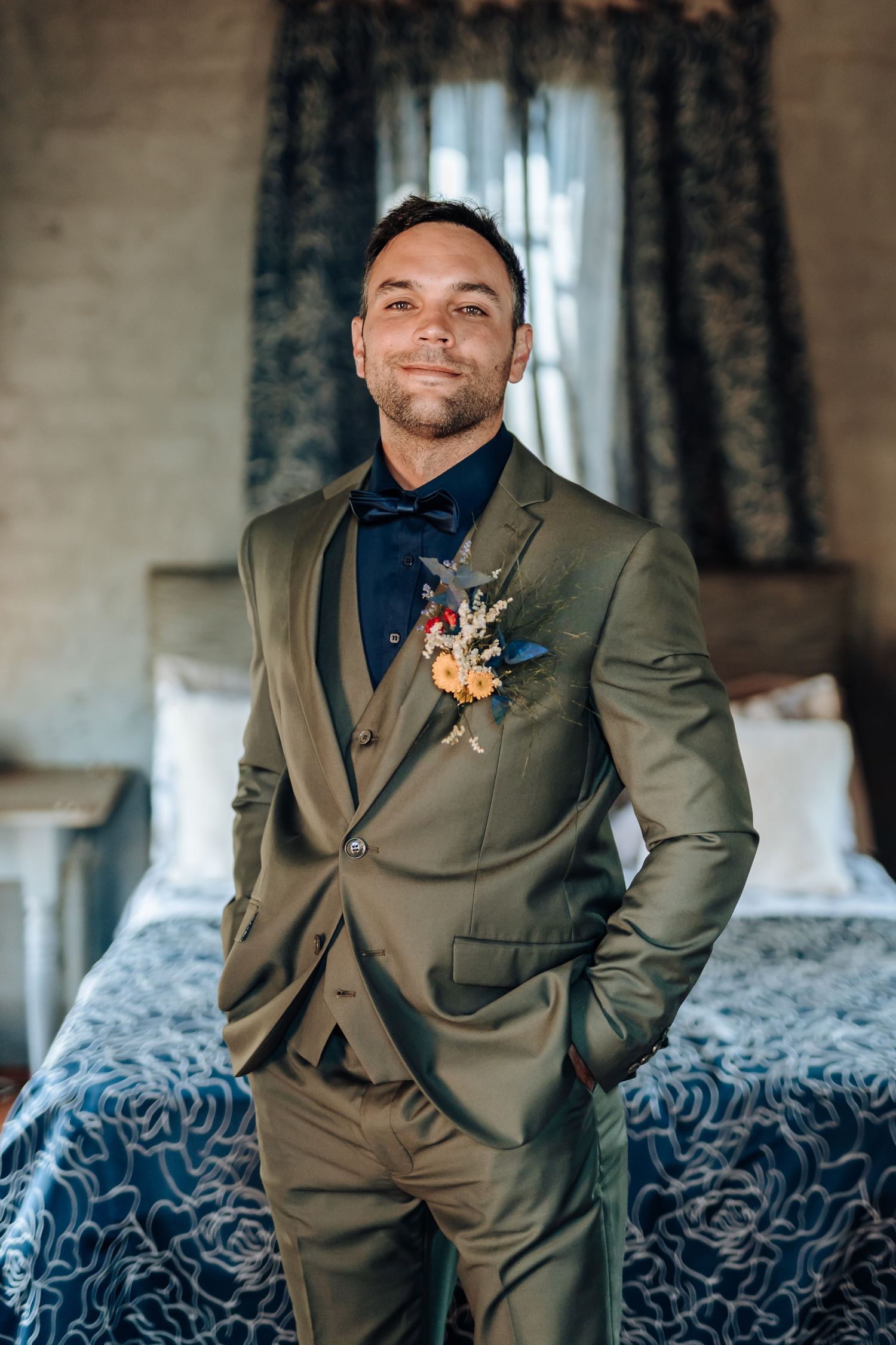 Person in olive suit with boutonniere poses confidently in bedroom setting.