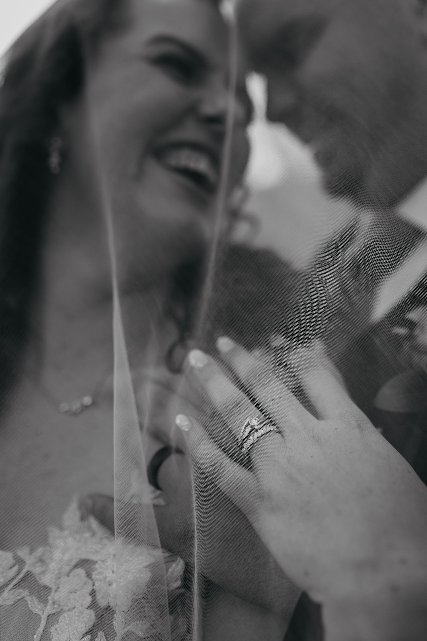 A close up black and white photograph showing a laughing bride displaying her wedding rings and lace dress details.