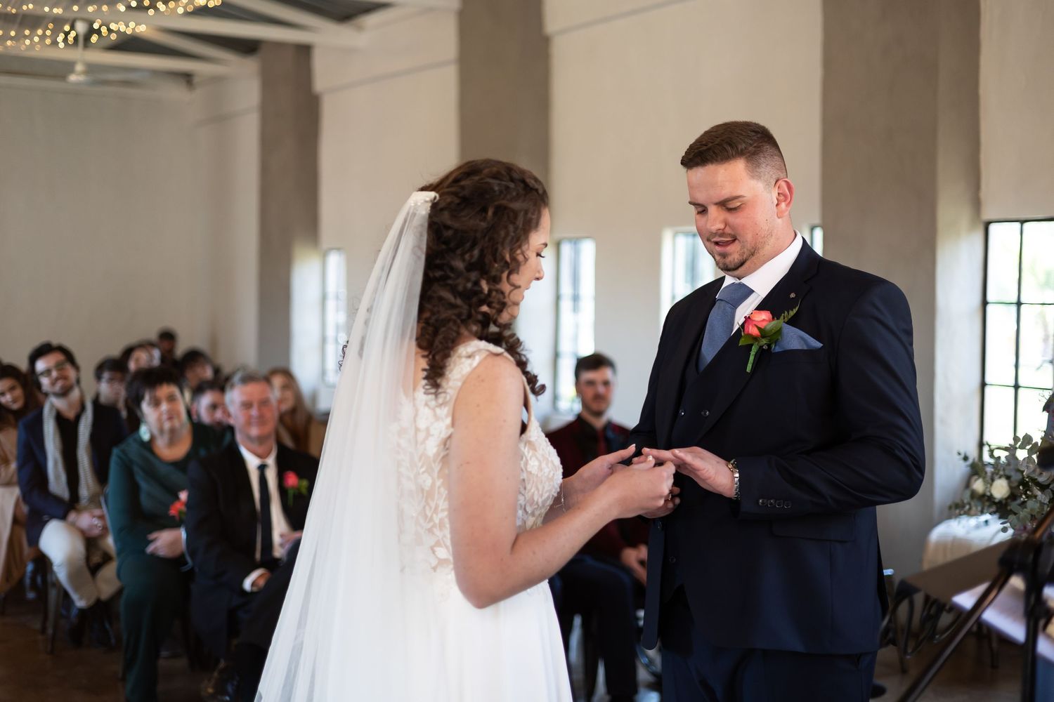 Two people exchange rings during a wedding ceremony in front of seated guests in an elegant indoor venue.