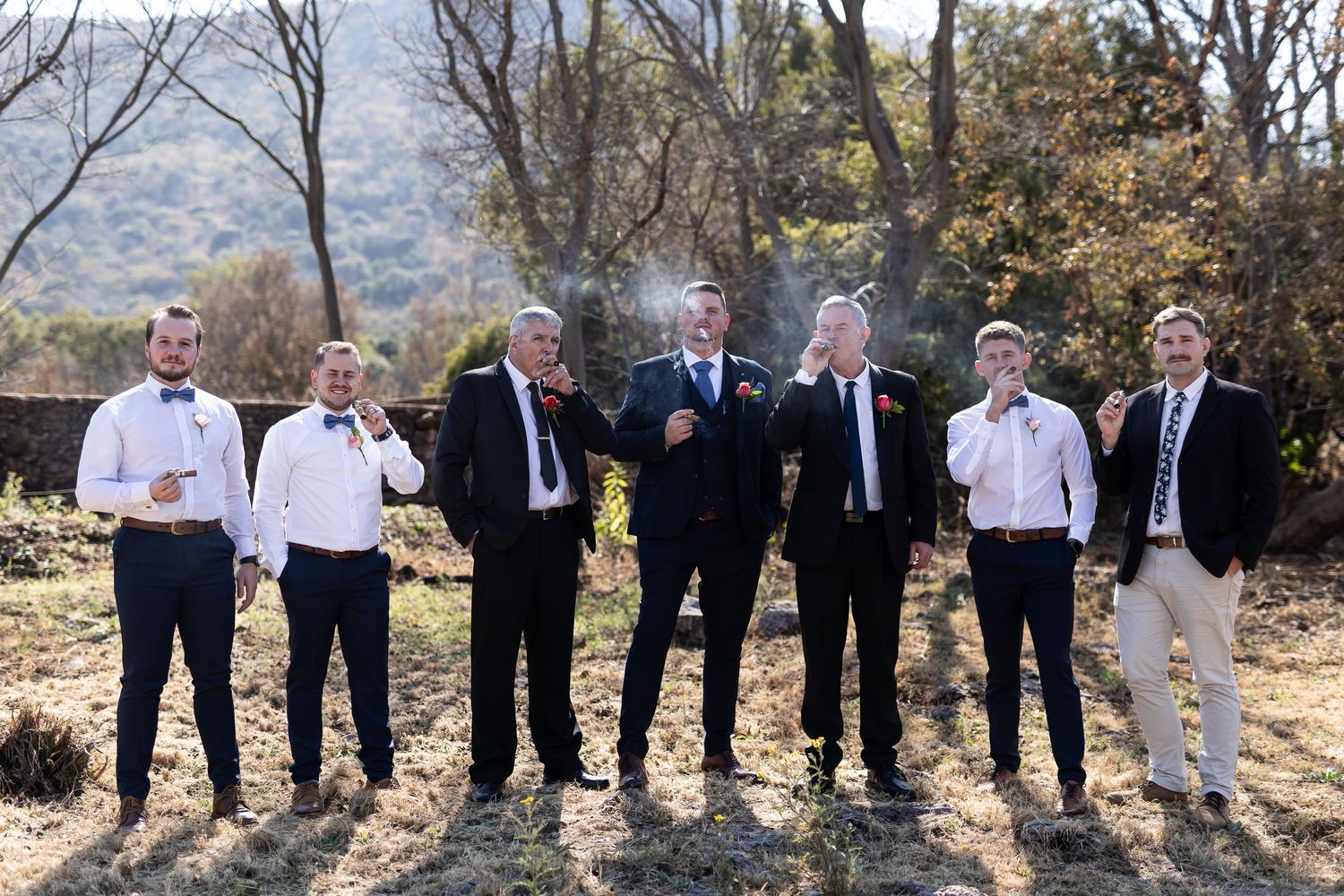 A casual group photo outdoors with multiple people standing in a row against a natural backdrop of trees and greenery.