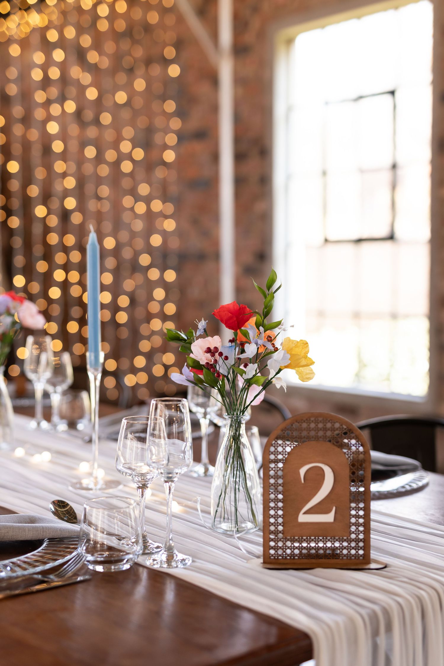 Rustic table setting with table number two, string lights, and scattered flower vases by brick wall windows.