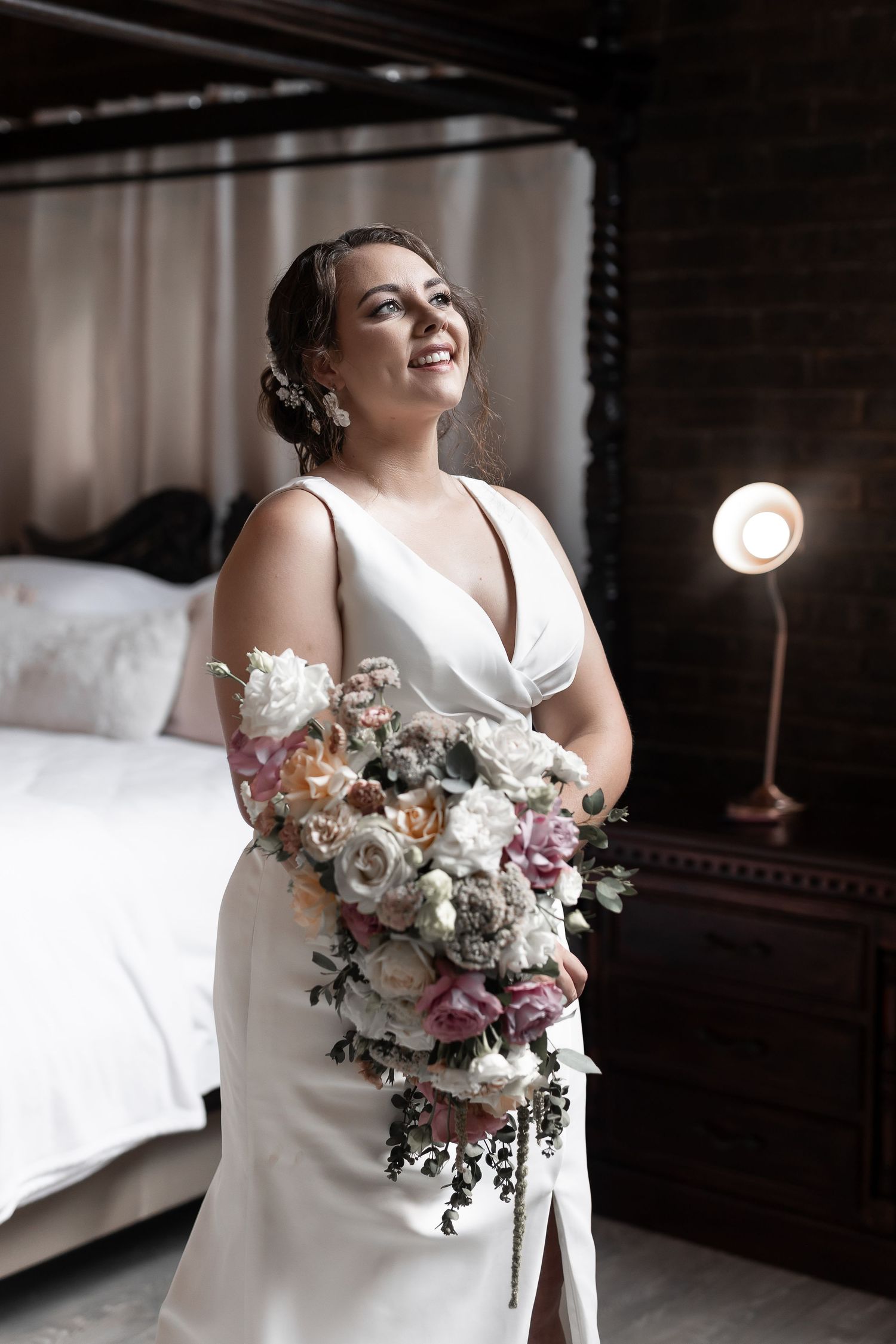 Bride holds lush floral bouquet while posing in classic white dress against bedroom backdrop.
