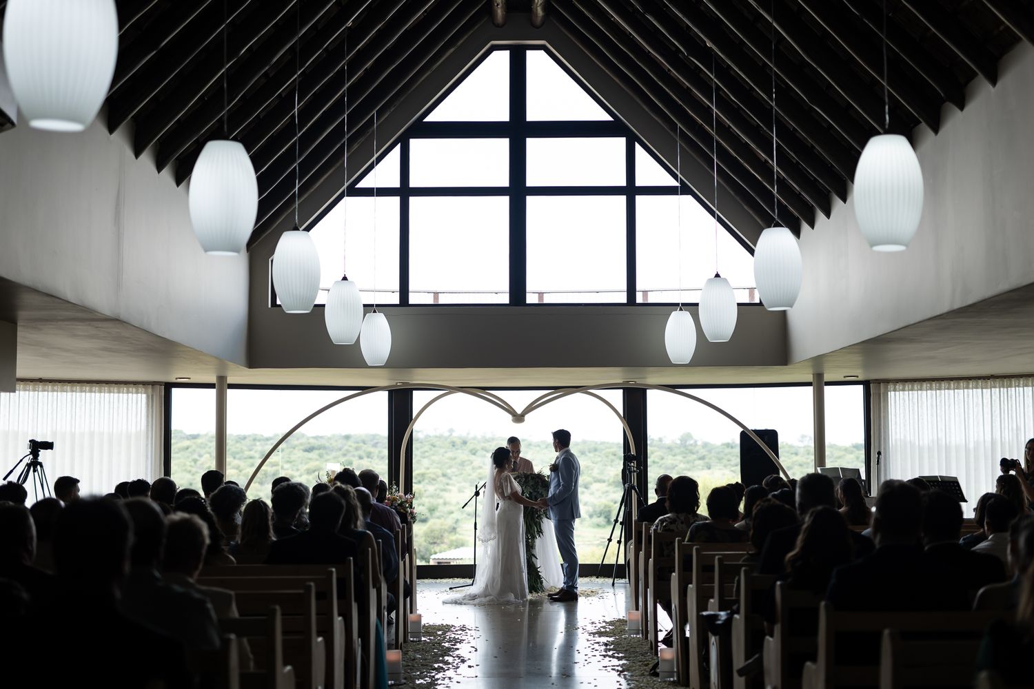 Silhouette of a wedding ceremony in a modern chapel with pendant lights and dramatic window backdrop.