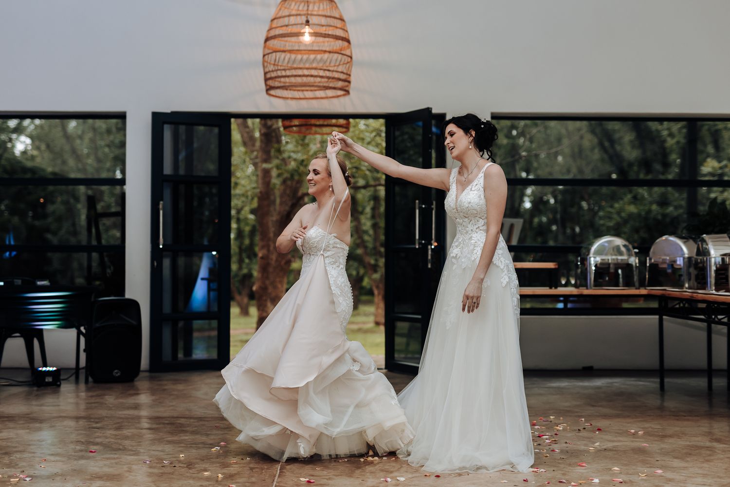 Two people in wedding dresses dance and twirl in a modern reception venue with rattan lighting.