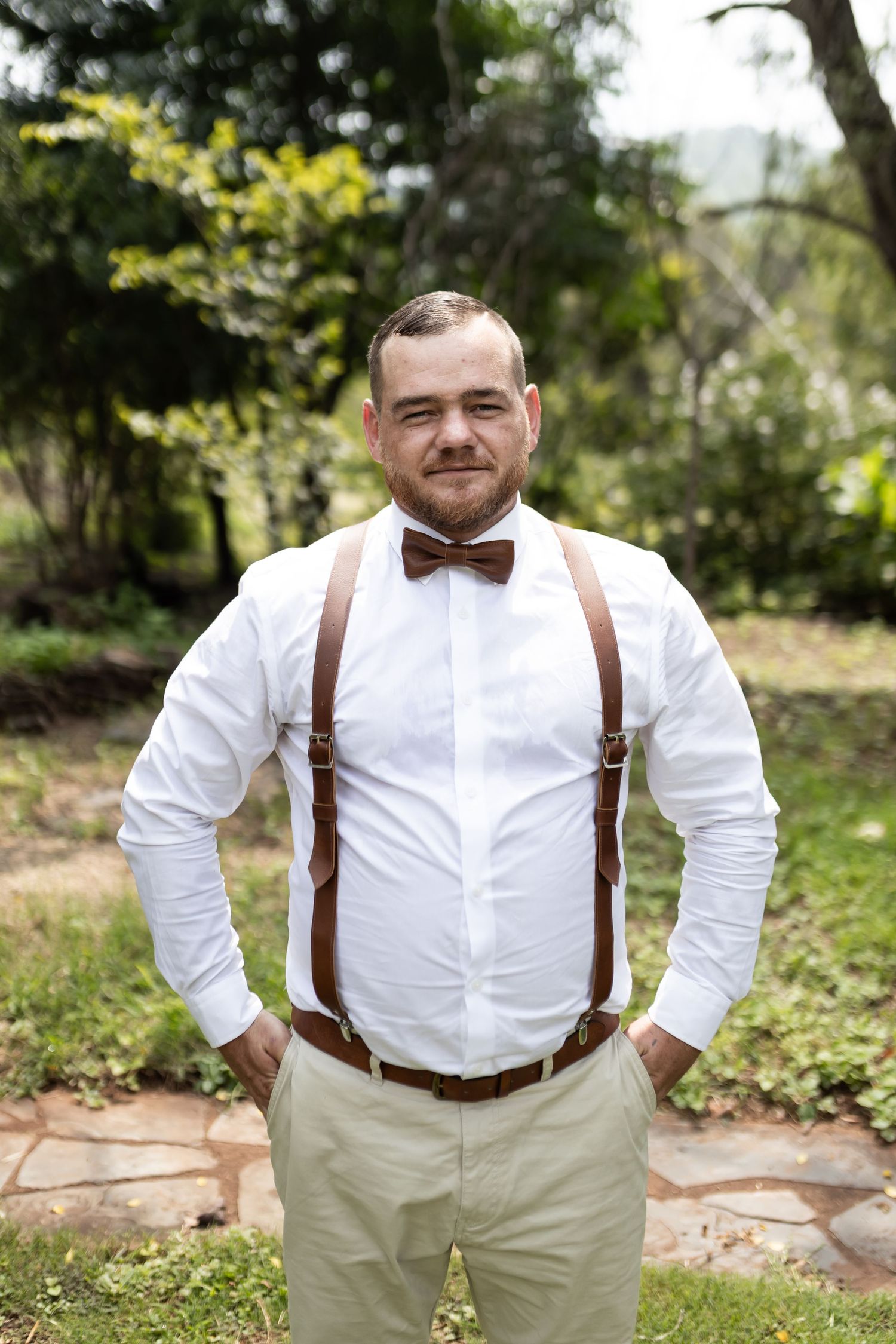 Groomsman in formal attire with brown suspenders and bow tie stands on stone path outdoors.