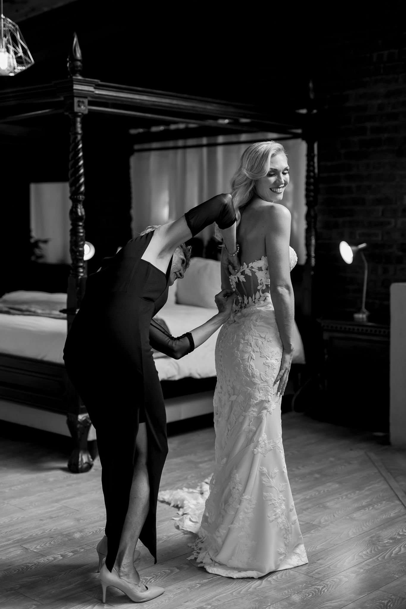 A bride in a fitted lace gown gets final adjustments in a room with a four-poster bed.
