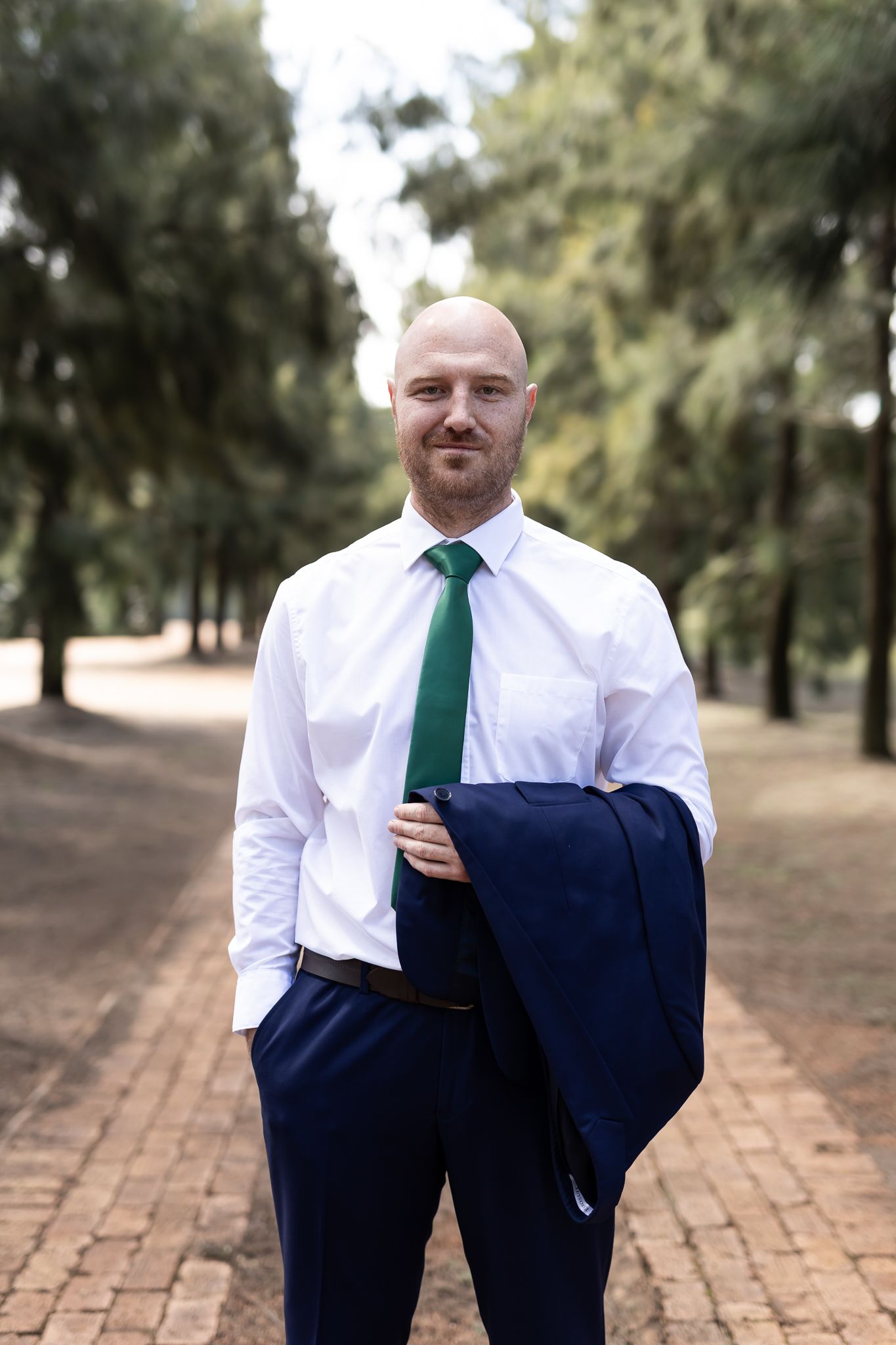 Groomsman in navy suit and emerald tie poses on brick path among pine trees.
