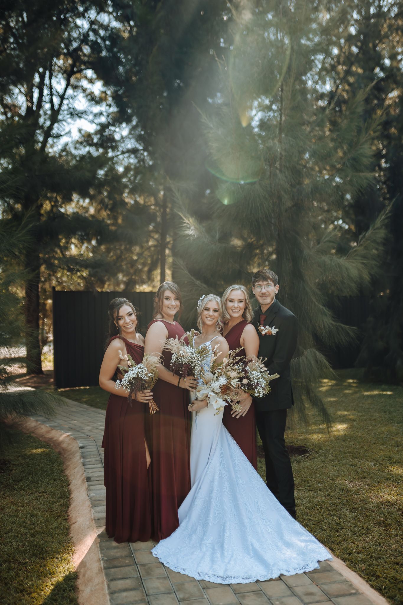 Wedding party in burgundy dresses poses on garden path with evergreen trees in background.