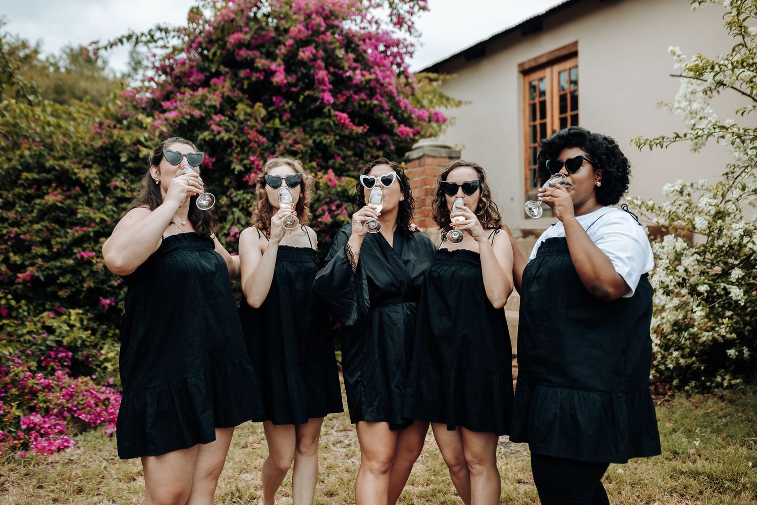 Wedding party in matching black dresses striking a funny pose while drinking from bottles in a garden with pink flowers.