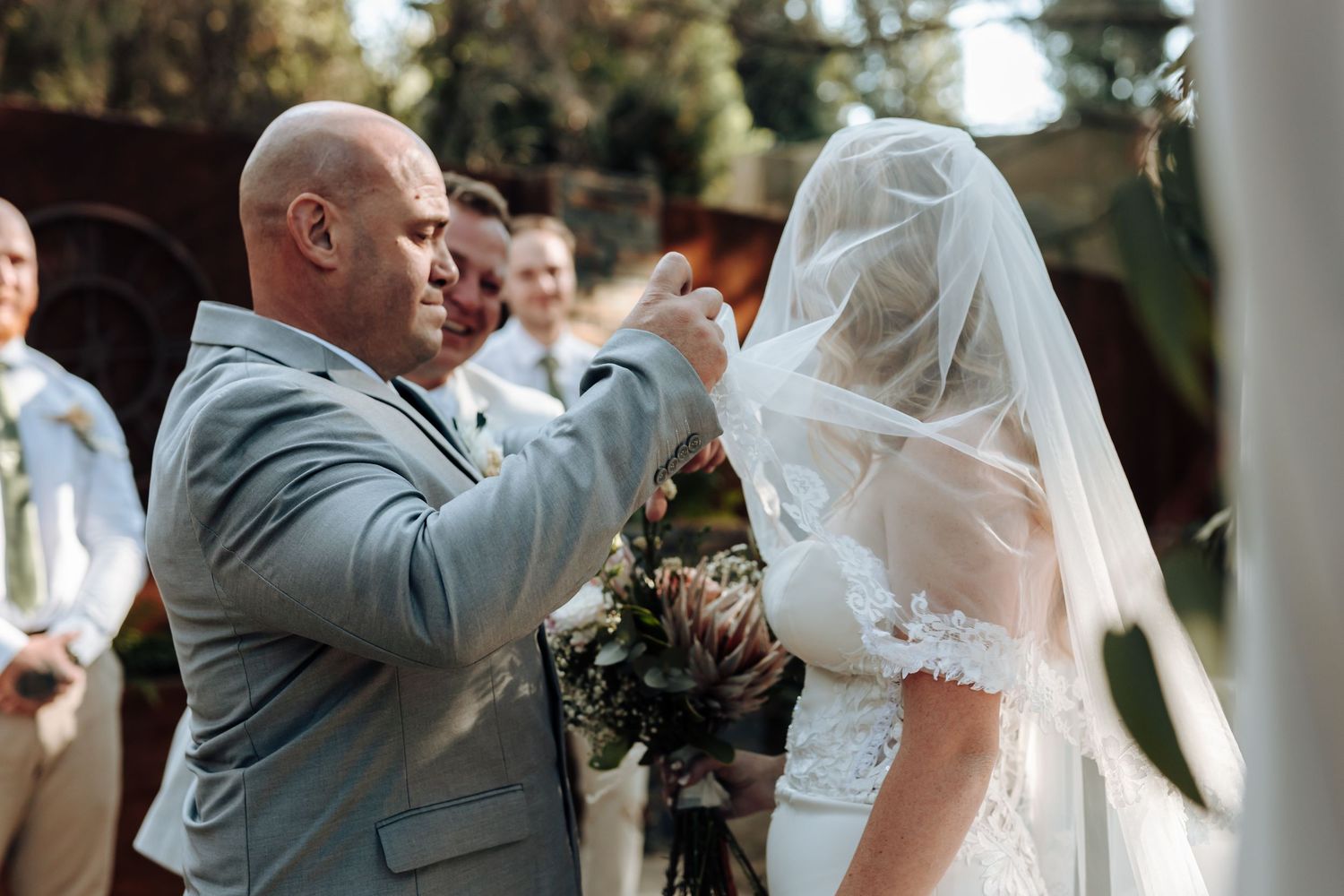 Wedding ceremony in a sunlit outdoor setting showing a tender moment of adjusting the bride's veil.