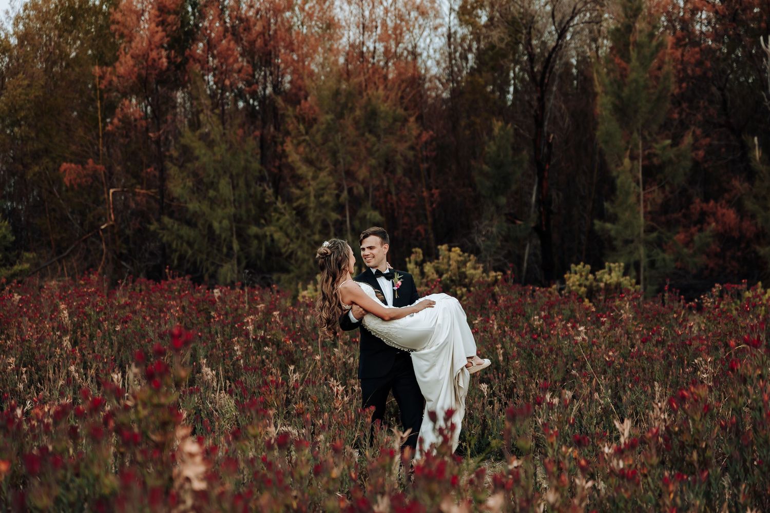 Groom carries bride in romantic pose among red wildflowers and autumn foliage.