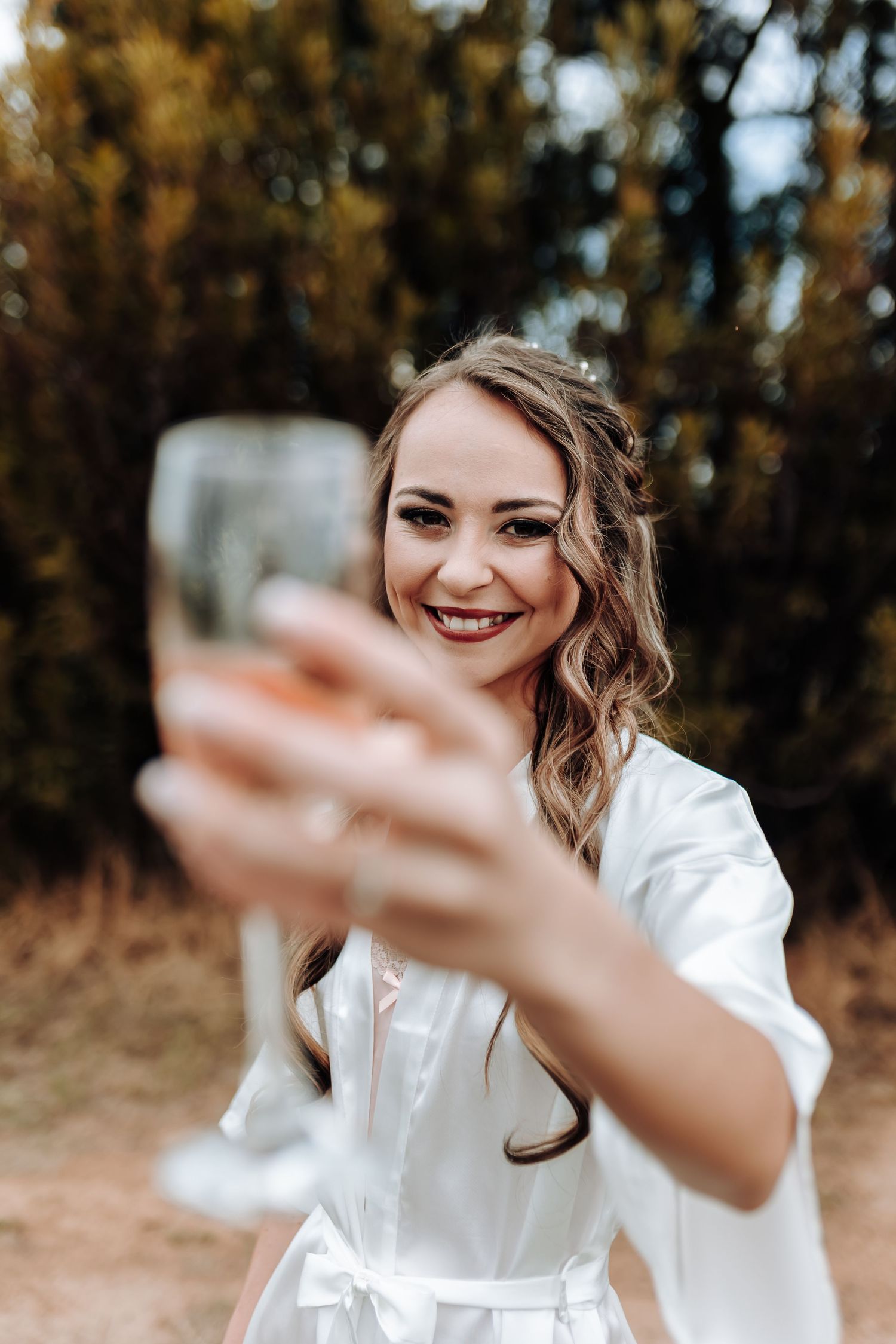 A person in a white robe holds up a champagne glass while smiling during pre-wedding celebrations.