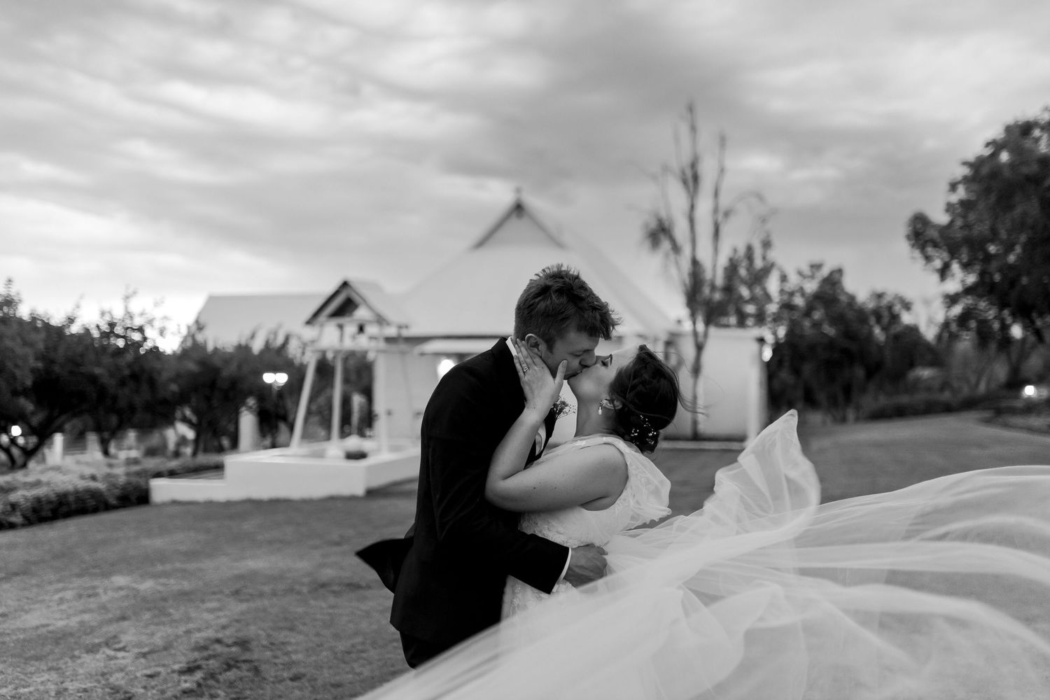 Black and white wedding photo showing an intimate moment between newlyweds with a chapel backdrop.