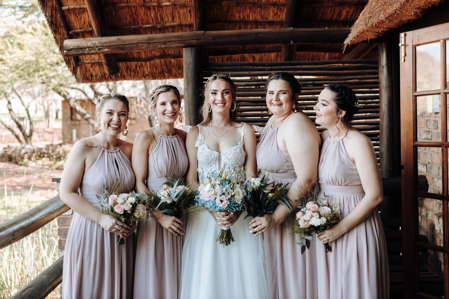 Bridal party photo with bridesmaids in matching dusty rose dresses under thatched roof.