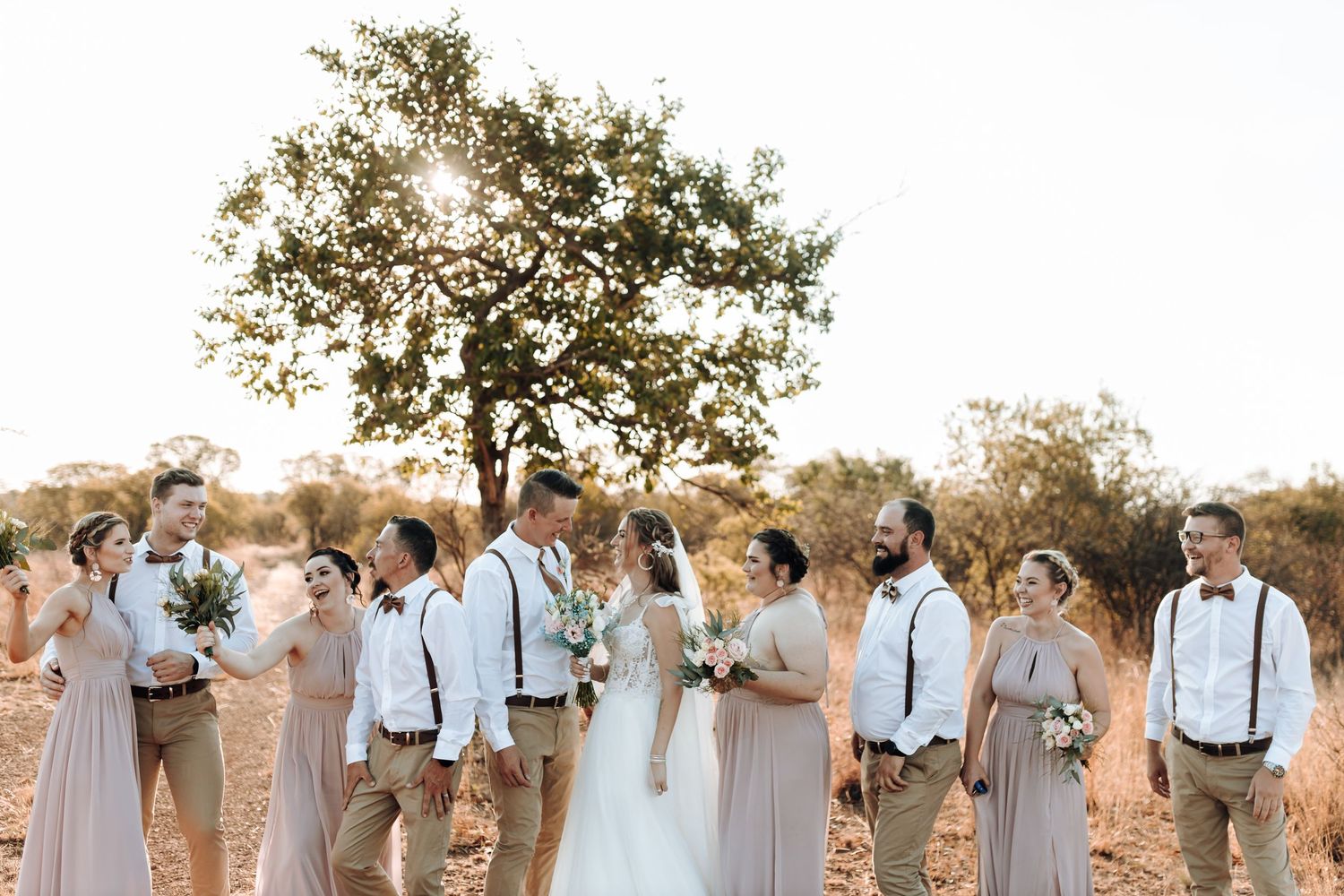 Wedding group laughs together under a tree in warm sunset lighting.
