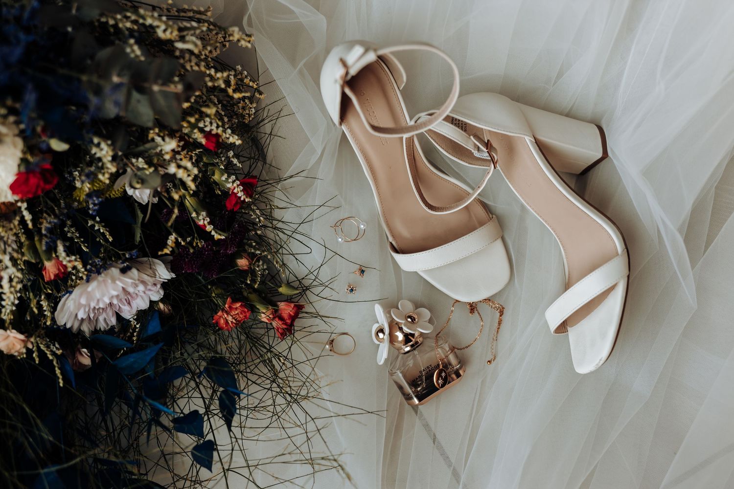 White wedding shoes and rings styled with delicate floral decor on tulle fabric.