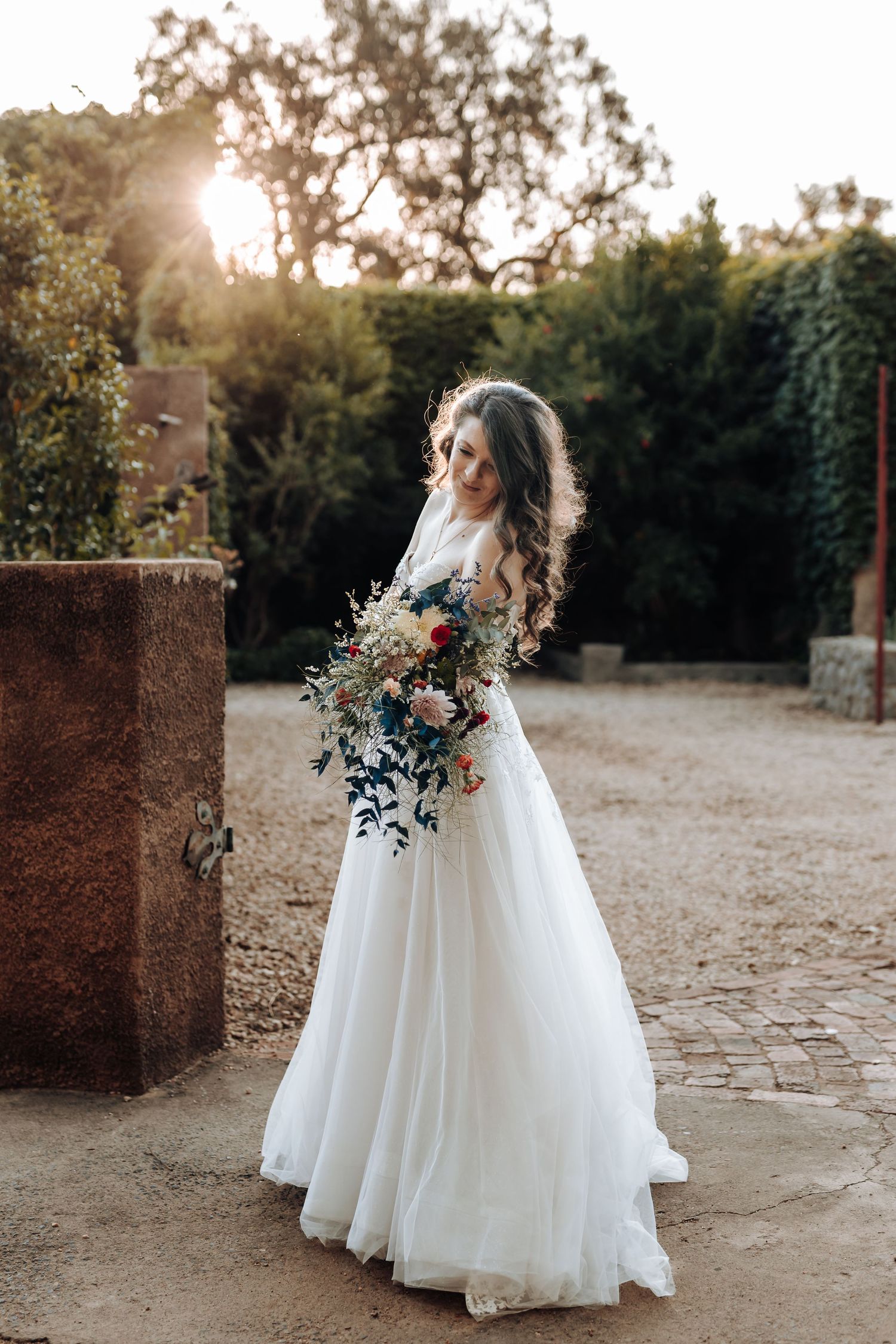 A bride in a flowing white gown holds a cascading bouquet while backlit by golden sunlight.