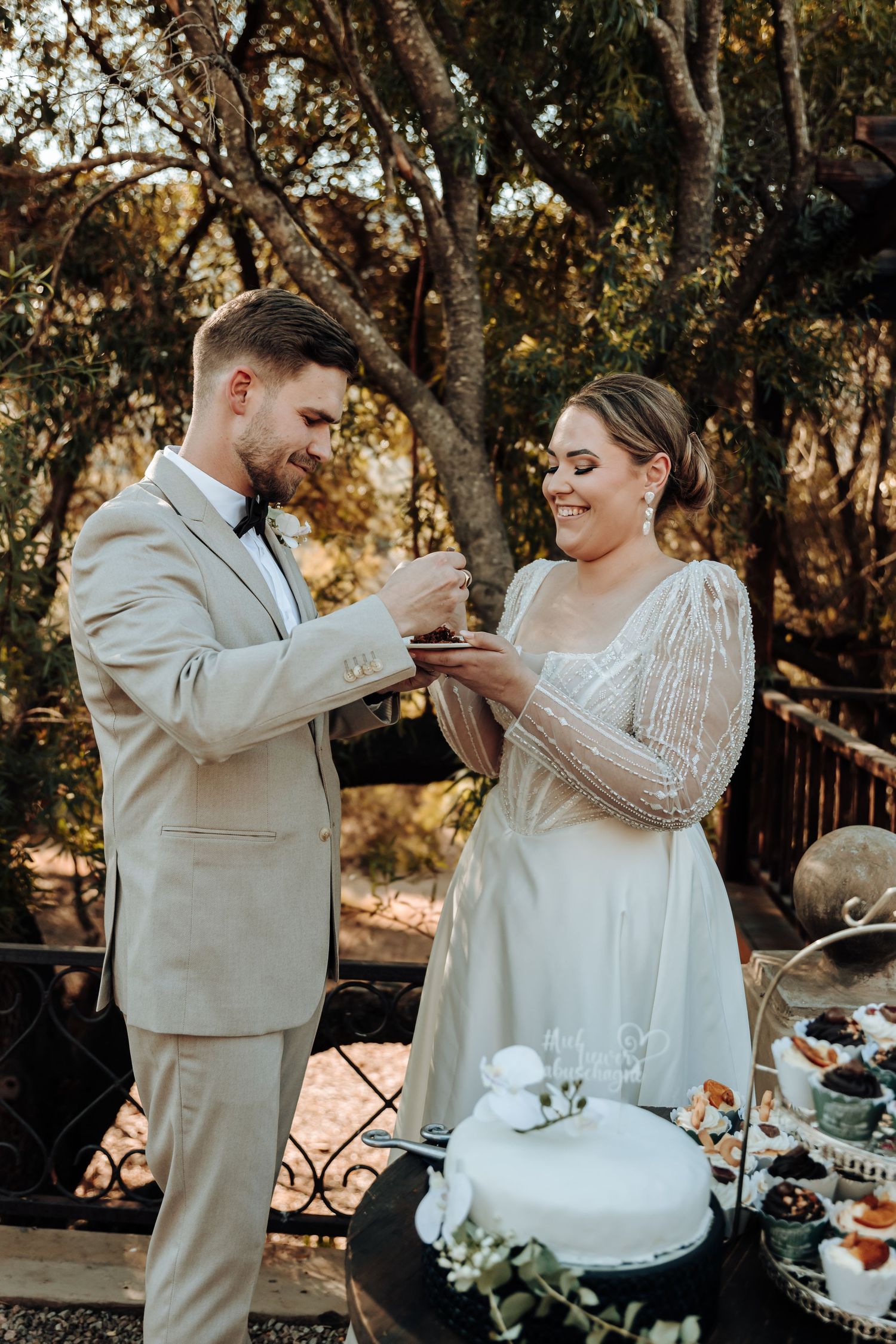 A couple wearing light-colored wedding attire shares dessert during their outdoor reception.