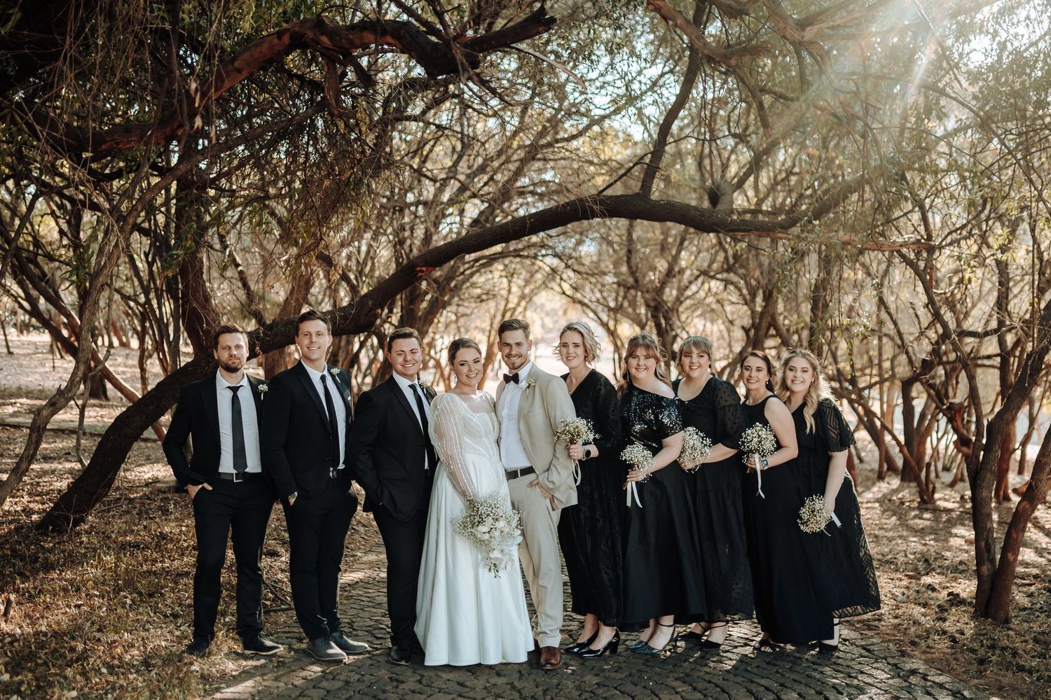 Wedding party poses together under an archway of trees in a rustic outdoor setting.
