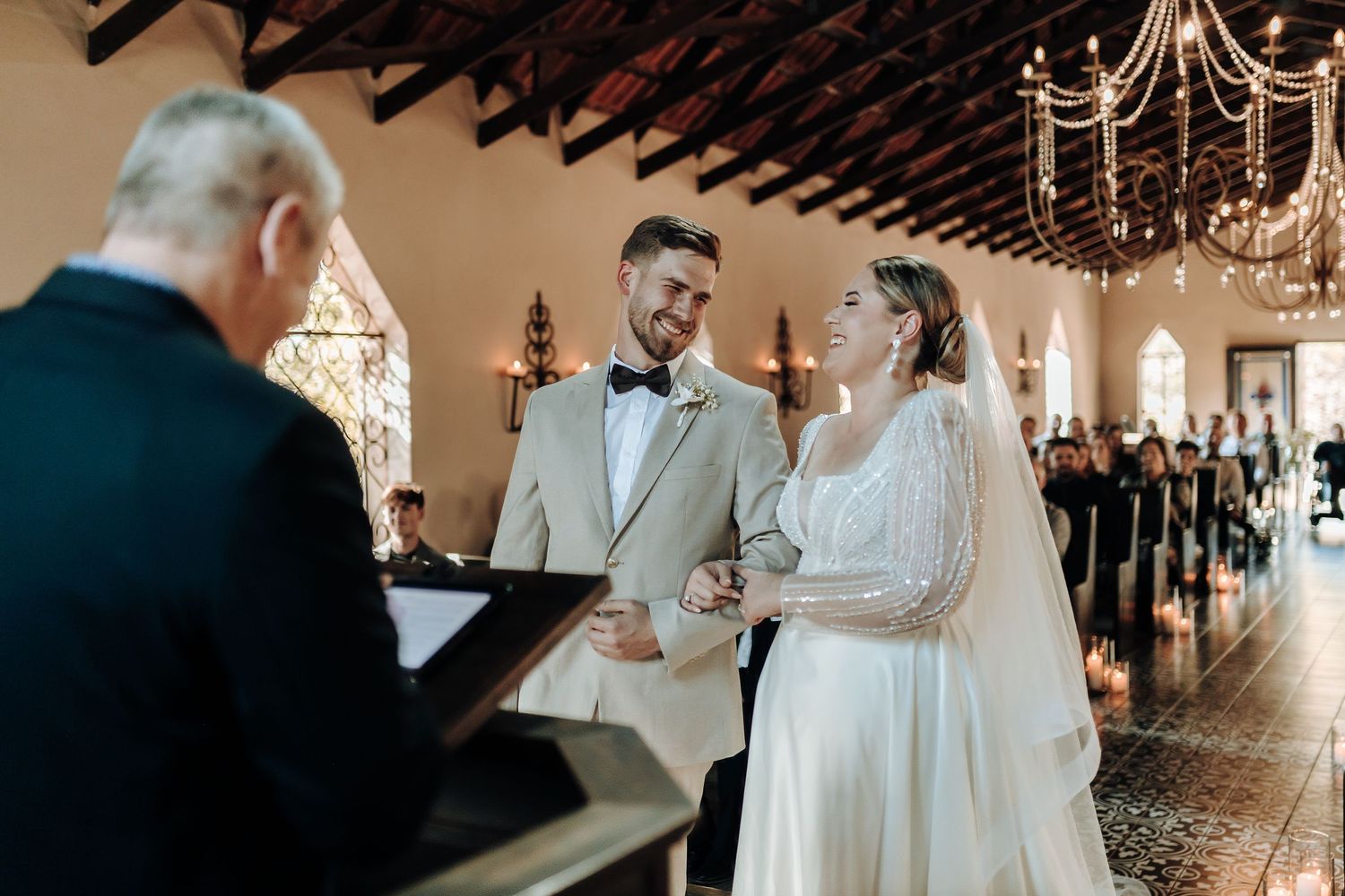 A wedding ceremony unfolds in a chapel with elegant chandeliers and stained glass windows.