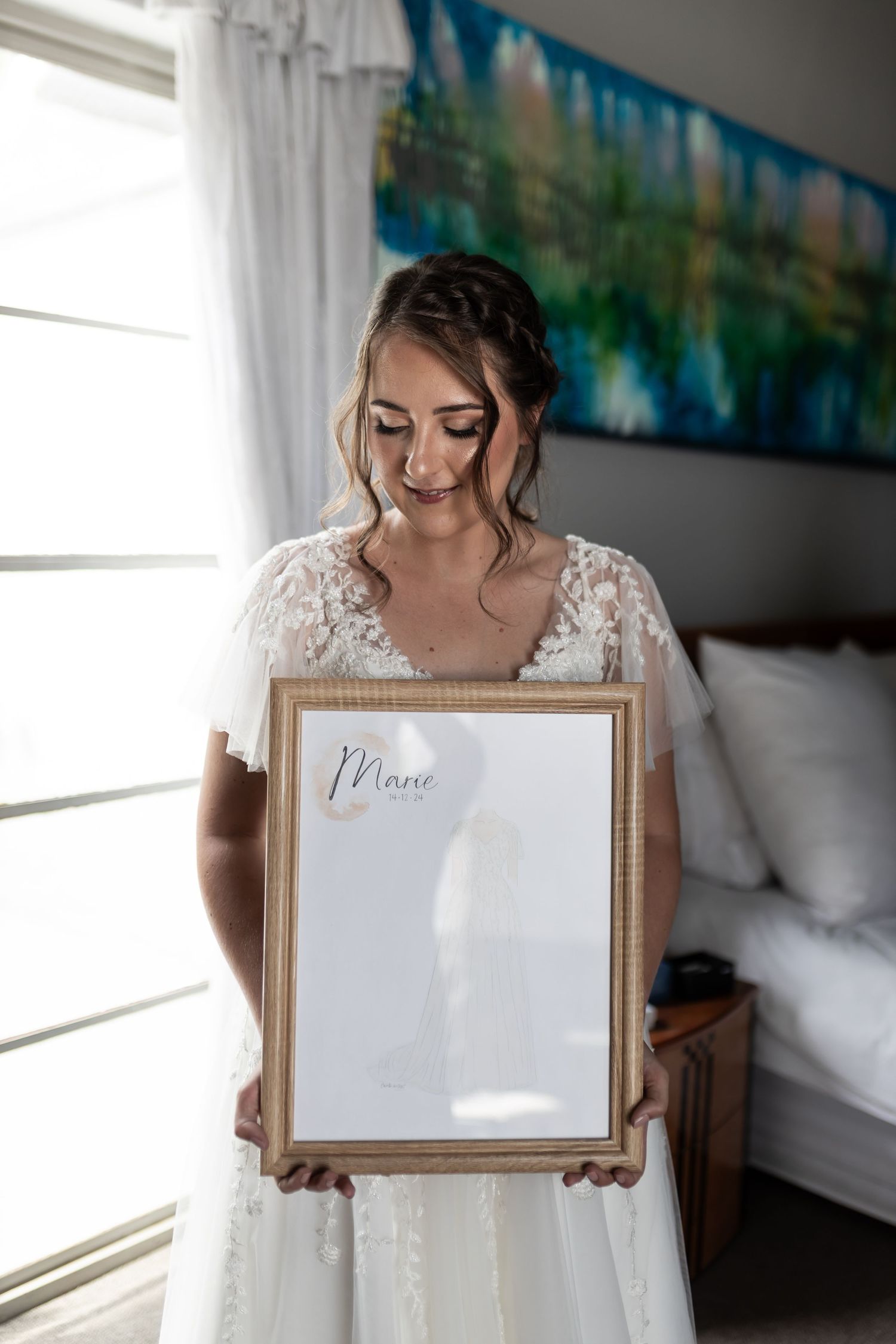 A person in a white lace wedding gown holds a framed artwork in a sunlit room.