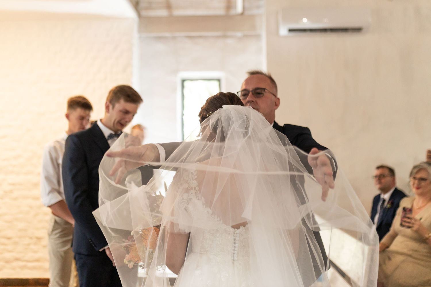 Emotional moment during wedding ceremony as veil is adjusted in bright chapel setting.