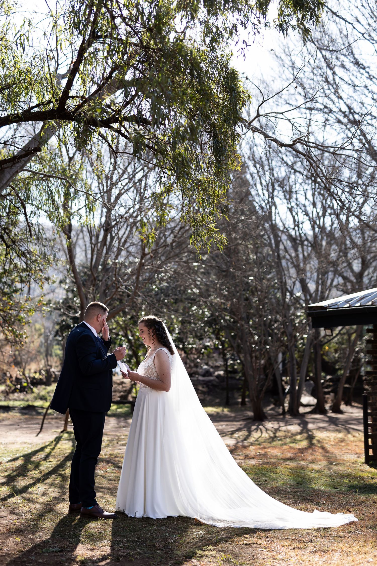 A bride and groom share a romantic moment in a wooded outdoor setting with sunlight filtering through the trees.