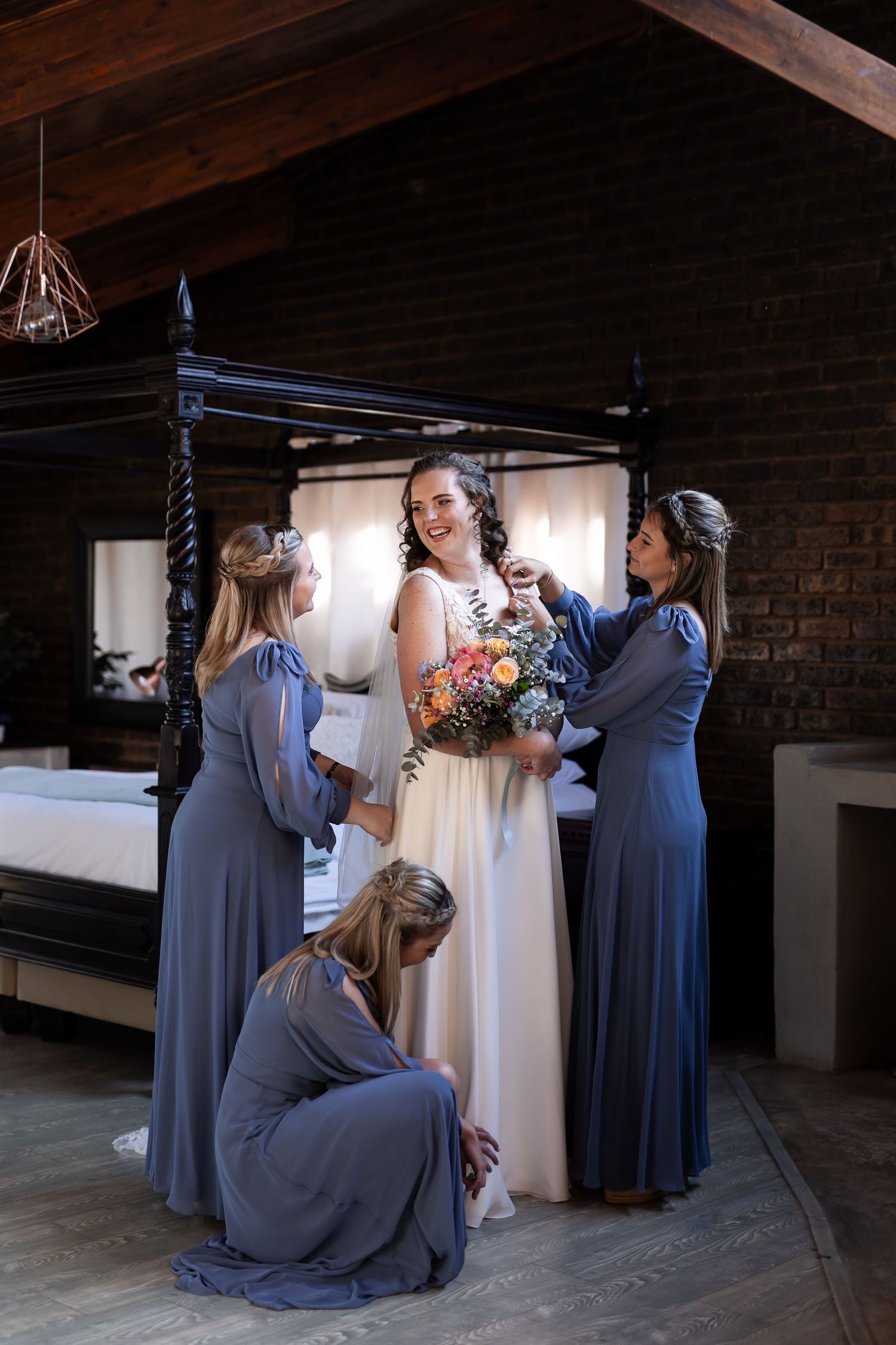 Wedding preparations with bridesmaids in dusty blue dresses helping adjust a white wedding gown in a rustic indoor setting.